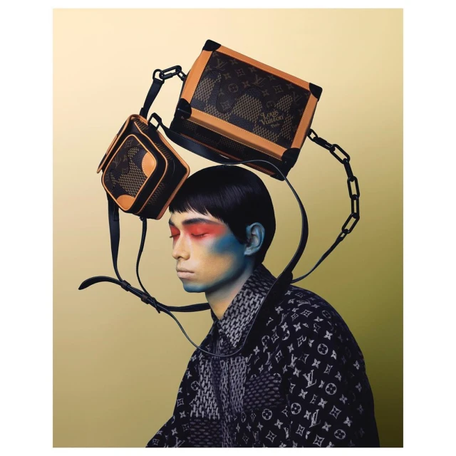 Editorial shot of a person with red and blue makeup, wearing a patterned jacket, and two black and tan bags floating above the head.
