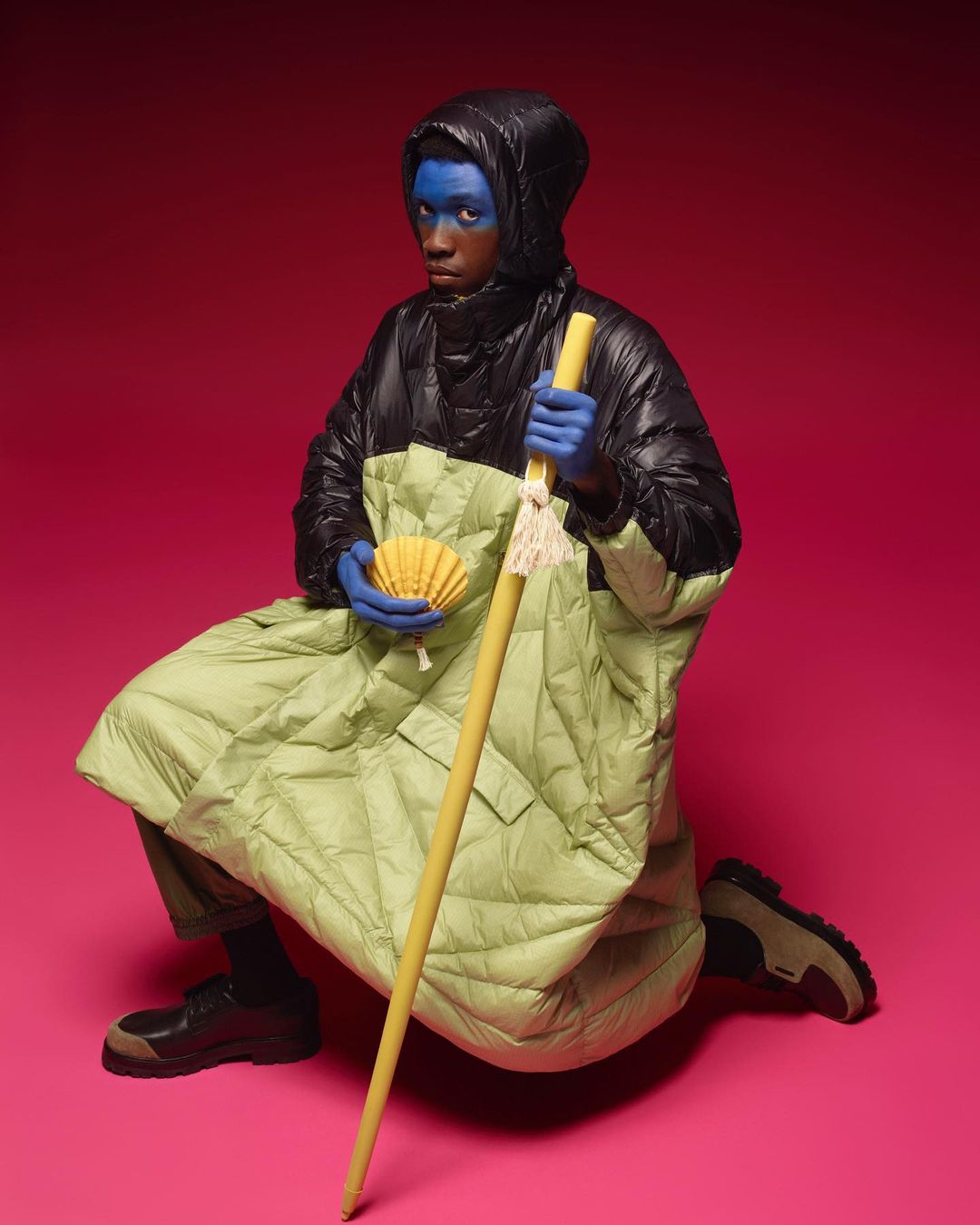 Person in black puffer jacket and lime green quilted poncho, wearing blue face paint, holding yellow staff and shell against pink backdrop.