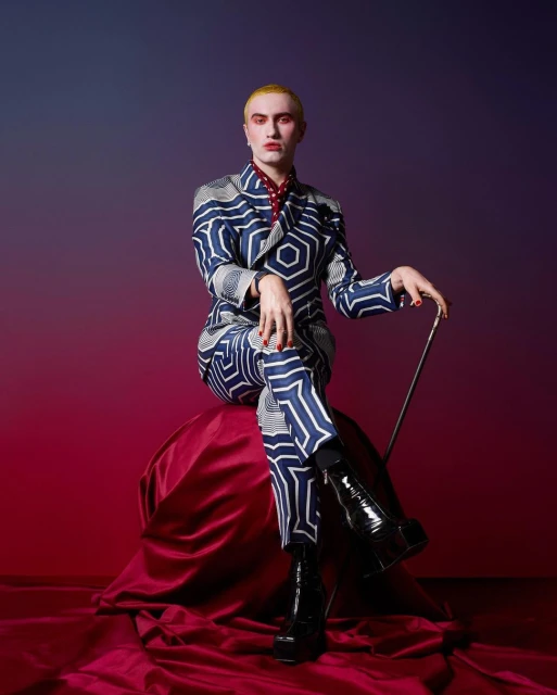 Person with blonde hair and patterned suit poses sitting on red draped fabric in a studio with red and purple gradient background.