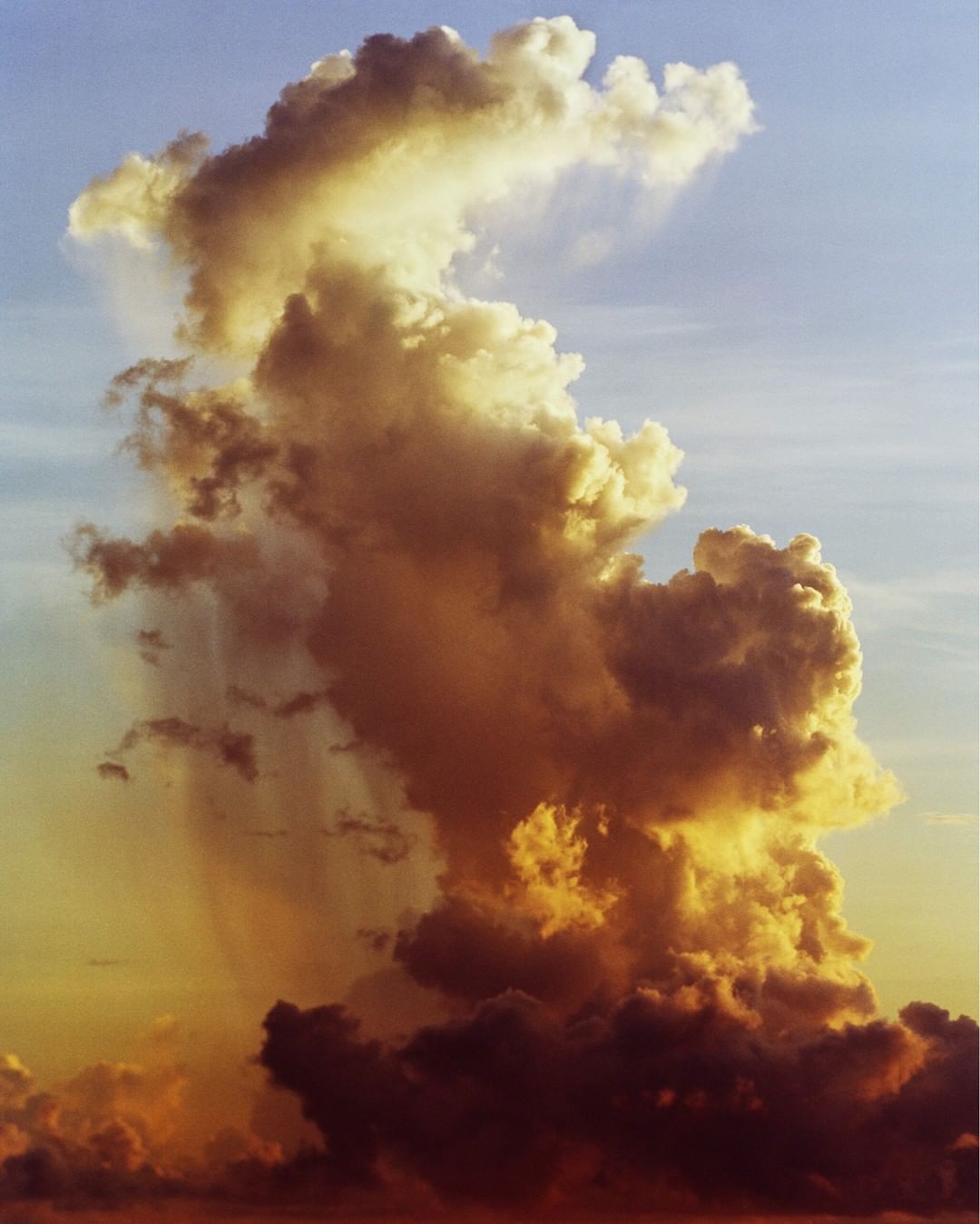 Dramatic cumulus cloudscape fills the frame with textured formations and vibrant golden light against a soft blue sky at daytime.