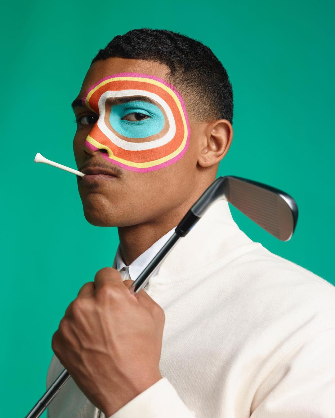 Close-up of a man with colorful face paint holding a golf club with a golf tee in his mouth against a green background.