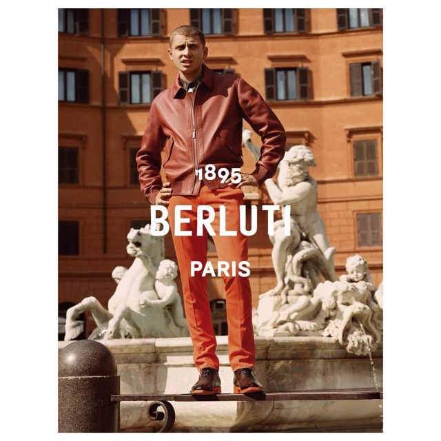 Man in leather jacket and orange trousers posing in front of a building and fountain in an outdoor fashion shot for Berluti.
