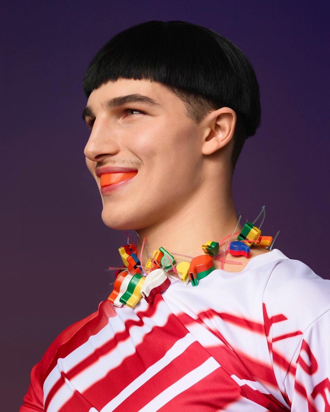 Stylish young man with bowl haircut smiles, wearing orange mouthguard and colorful necklace in a studio portrait against a purple backdrop.