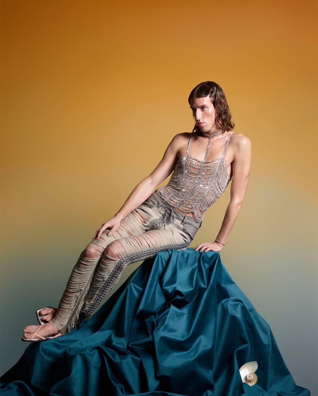 A single male fashion model poses while seated on draped fabric, wearing a silver top and distressed denim pants, with a gold background.