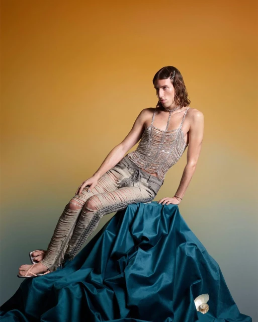 A single male fashion model poses while seated on draped fabric, wearing a silver top and distressed denim pants, with a gold background.
