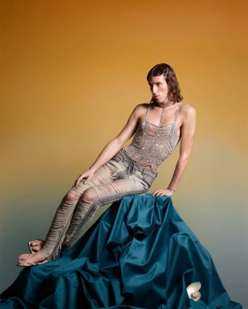 A single male fashion model poses while seated on draped fabric, wearing a silver top and distressed denim pants, with a gold background.
