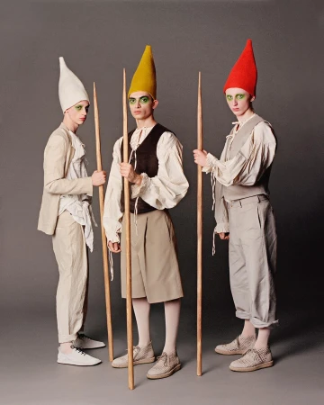 Three slender figures in costume stand in a photo studio. The costumes feature pointed hats, light fabrics, and theatrical makeup.