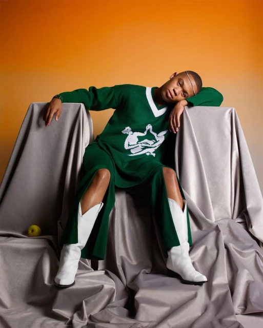 A person poses in a green outfit and white boots, seated on silver fabric with orange background. Studio lighting enhances the shot.