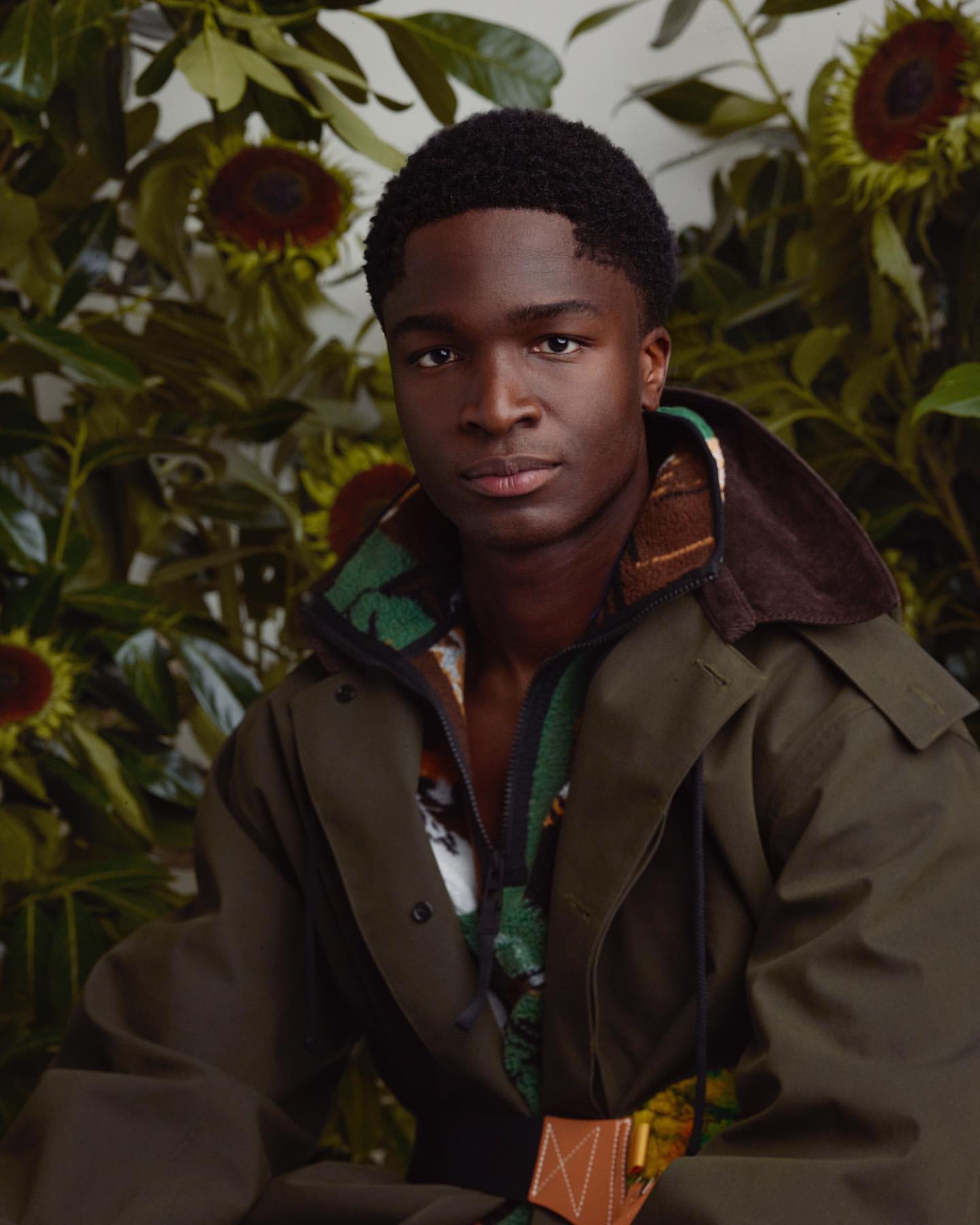 Portrait of an African American male model with a short haircut, wearing a green jacket, posed in front of a sunflower and foliage backdrop.