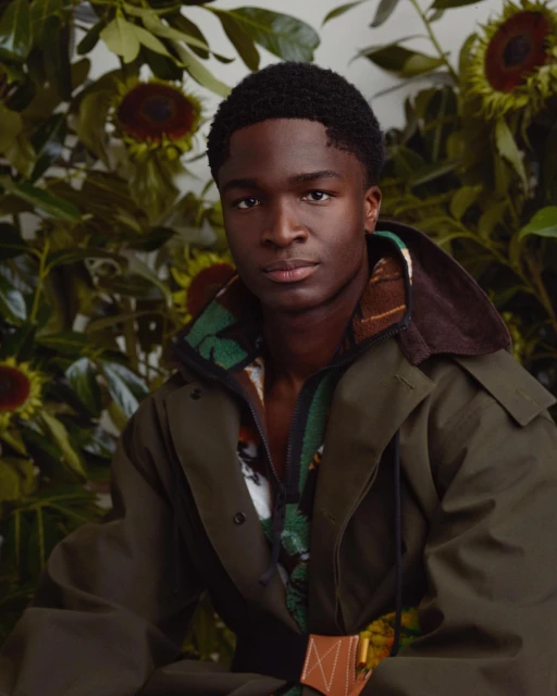 Portrait of an African American male model with a short haircut, wearing a green jacket, posed in front of a sunflower and foliage backdrop.
