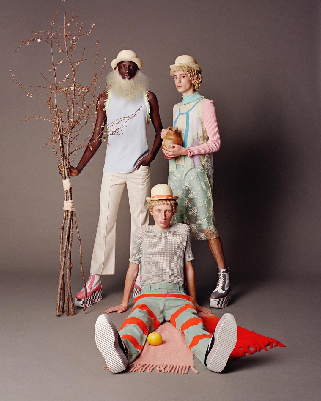 Three fashion models pose indoors in hats and outfits with unique textures, colors, and platform shoes against a plain grey background.