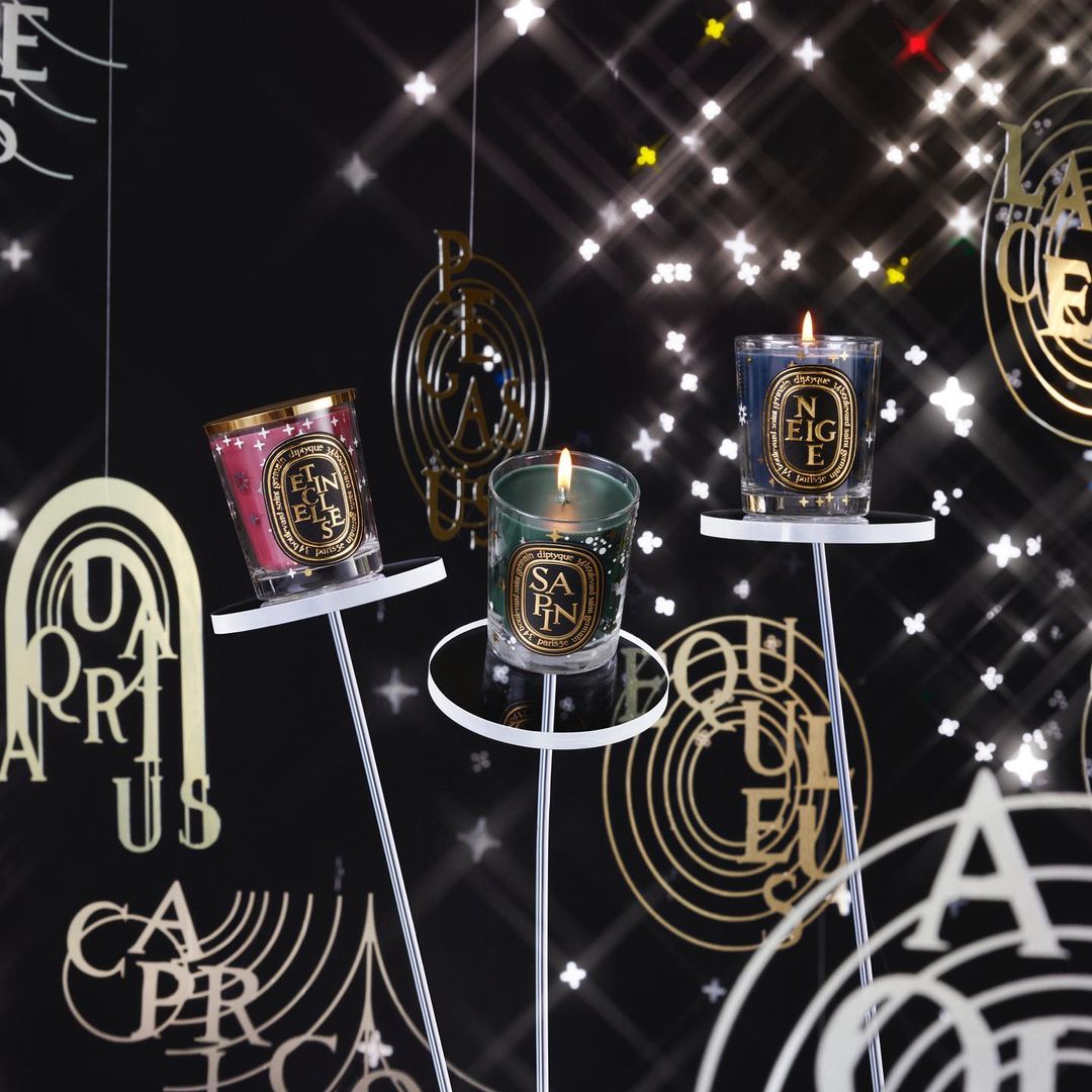 Three Diptyque candles displayed on stands against a backdrop of ornate gold lettering and sparkling stars, creating a festive ambiance.