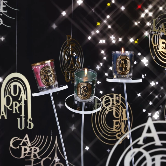 Three Diptyque candles displayed on stands against a backdrop of ornate gold lettering and sparkling stars, creating a festive ambiance.
