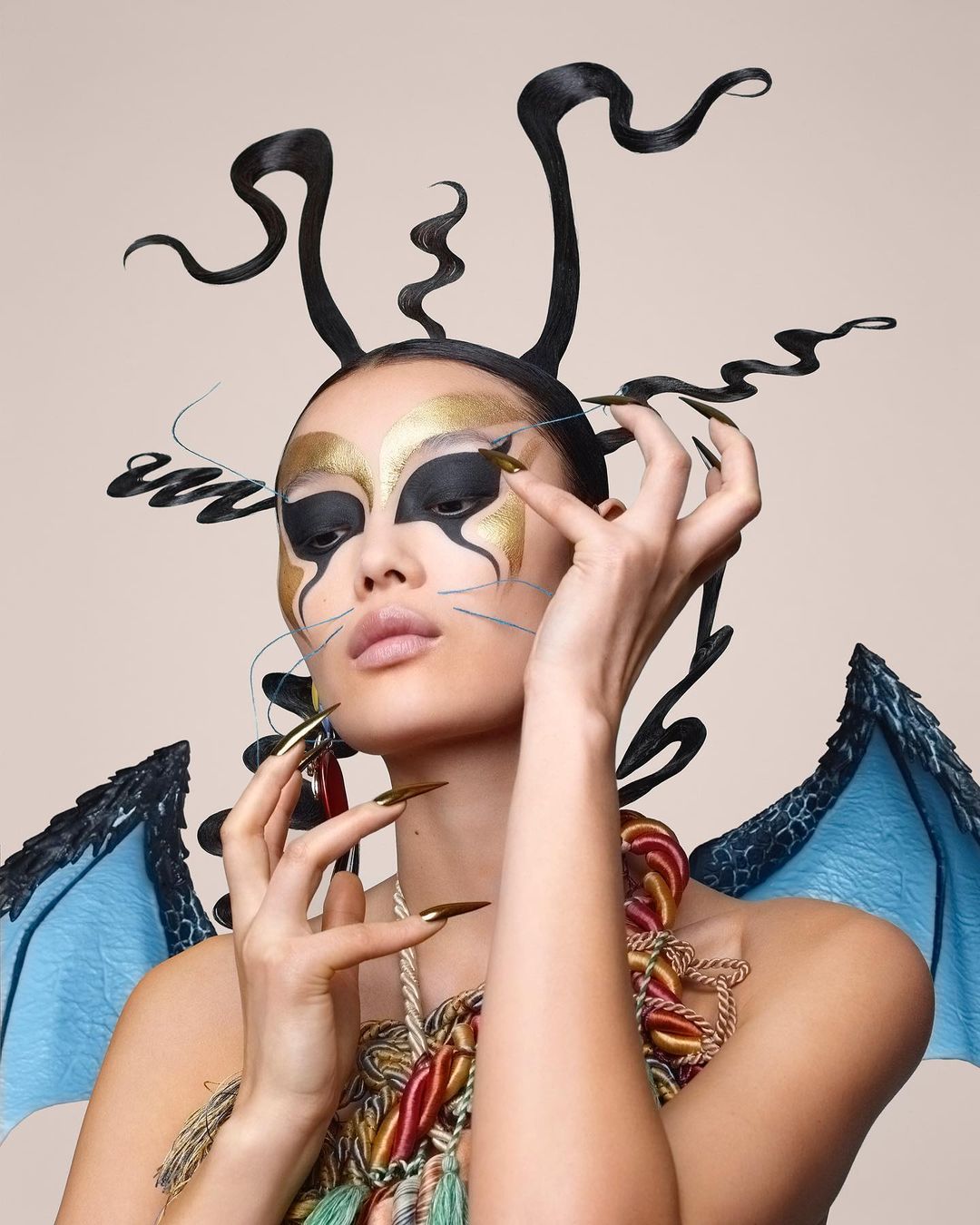 Woman with dramatic black and gold makeup and elaborate hair styling poses in front of a neutral background in a beauty shot.