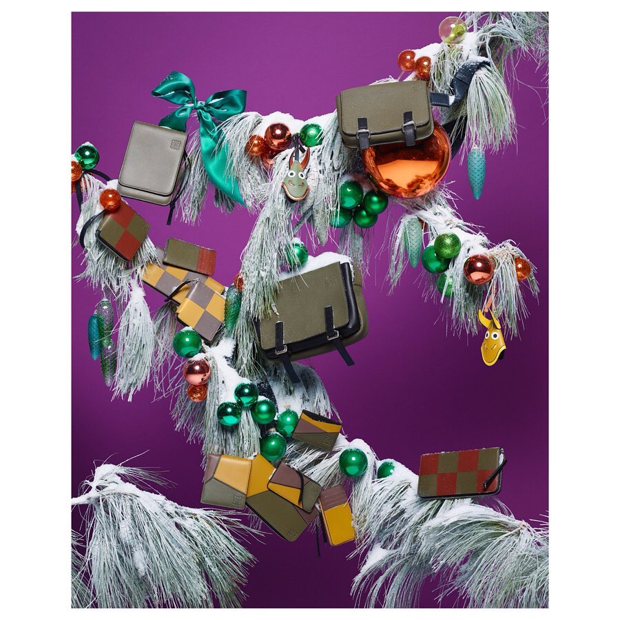 Luxury handbags and wallets displayed on a snow-covered pine branch with colorful Christmas ornaments against a purple background.