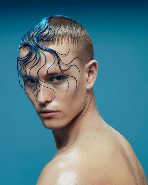 Close-up of a shirtless person with dyed blue hair styled in wavy strands across the forehead, posing against a blue backdrop in studio lighting.