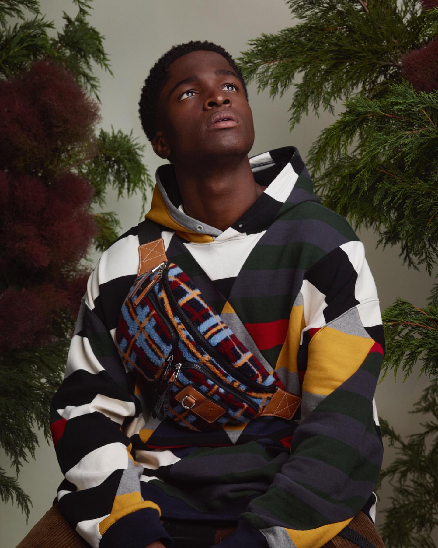 Young adult man wearing patterned hoodie and fanny pack, looking up, surrounded by foliage in a studio setting with neutral lighting.