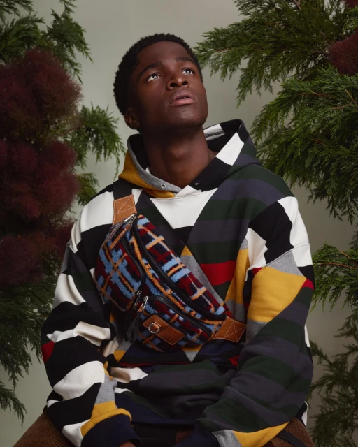Young adult man wearing patterned hoodie and fanny pack, looking up, surrounded by foliage in a studio setting with neutral lighting.