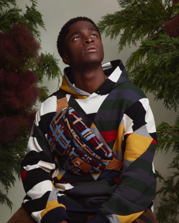 Young adult man wearing patterned hoodie and fanny pack, looking up, surrounded by foliage in a studio setting with neutral lighting.
