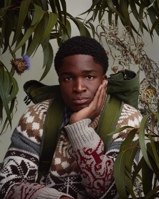 Portrait of a young Black man wearing a patterned sweater and green backpack, surrounded by green leaves and thistle flowers.