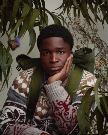 Portrait of a young Black man wearing a patterned sweater and green backpack, surrounded by green leaves and thistle flowers.