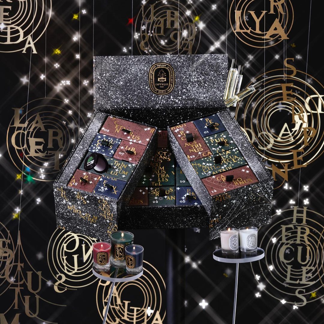Close-up of Diptyque advent calendar, green and white candles on stands, and gold letters suspended from the ceiling against black background.