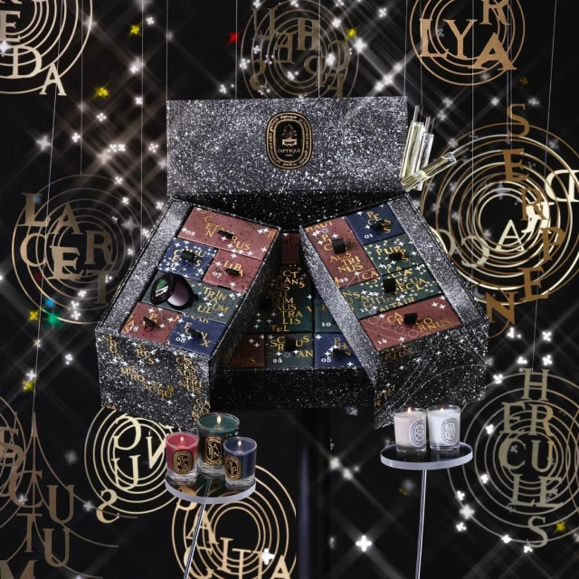 Close-up of Diptyque advent calendar, green and white candles on stands, and gold letters suspended from the ceiling against black background.