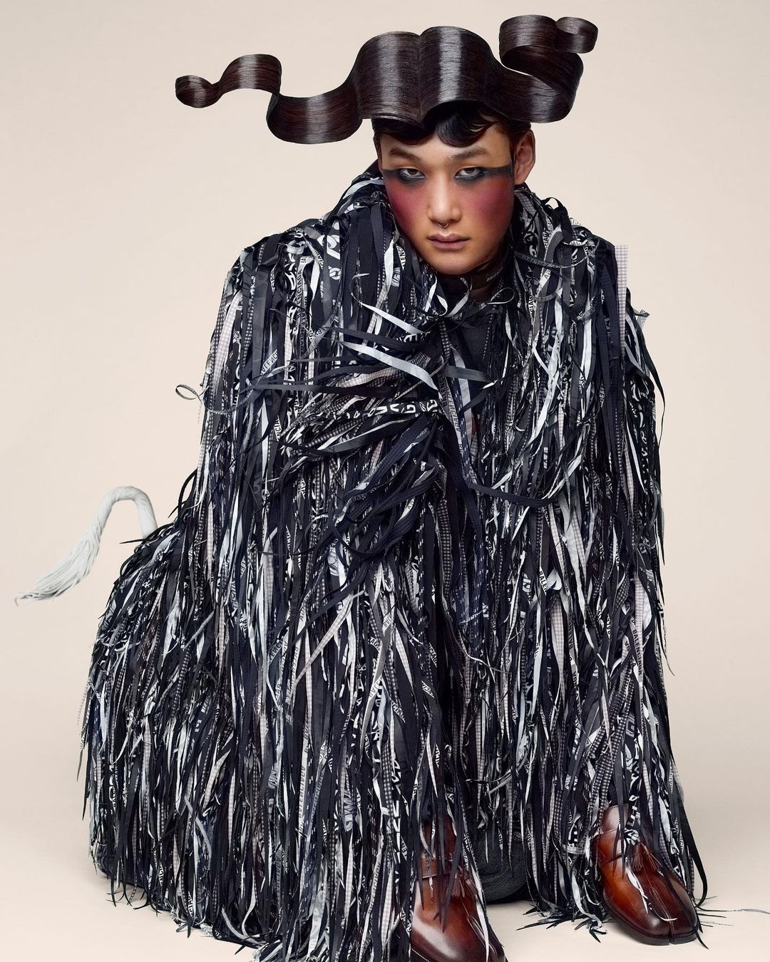 Asian adult male poses in an avant-garde fashion creation featuring a ribbon-fringed coat, unique hair styling, and bold makeup.