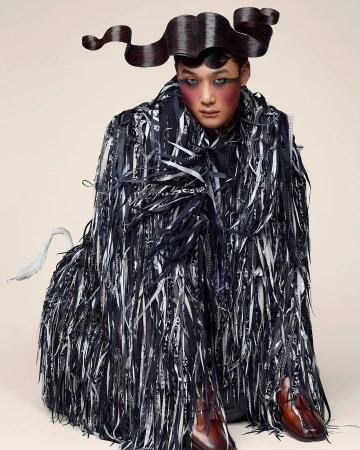 Asian adult male poses in an avant-garde fashion creation featuring a ribbon-fringed coat, unique hair styling, and bold makeup.