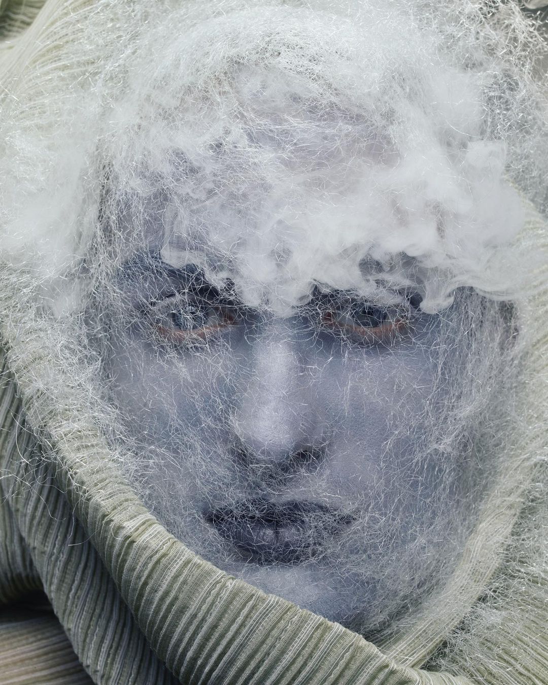 Portrait of a woman with a light blue face, wrapped in textured fabric and covered by a sheer, web-like material.