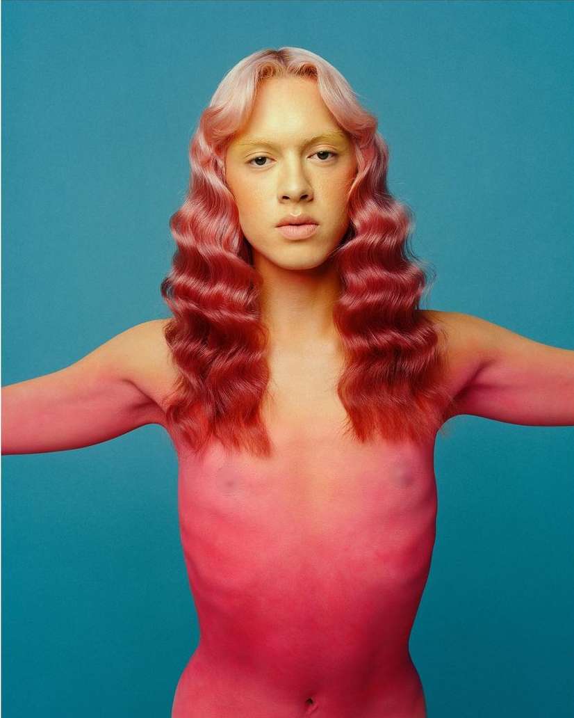 Portrait of androgynous human with pink skin, pink and blonde wavy hair, and nude body against turquoise background in a photo studio.