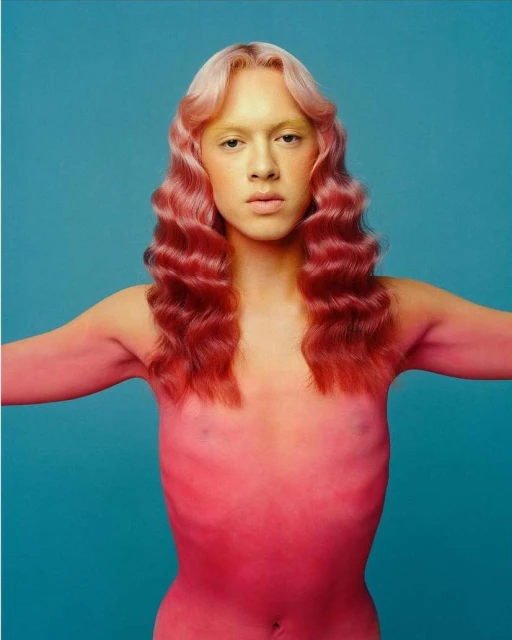 Portrait of androgynous human with pink skin, pink and blonde wavy hair, and nude body against turquoise background in a photo studio.