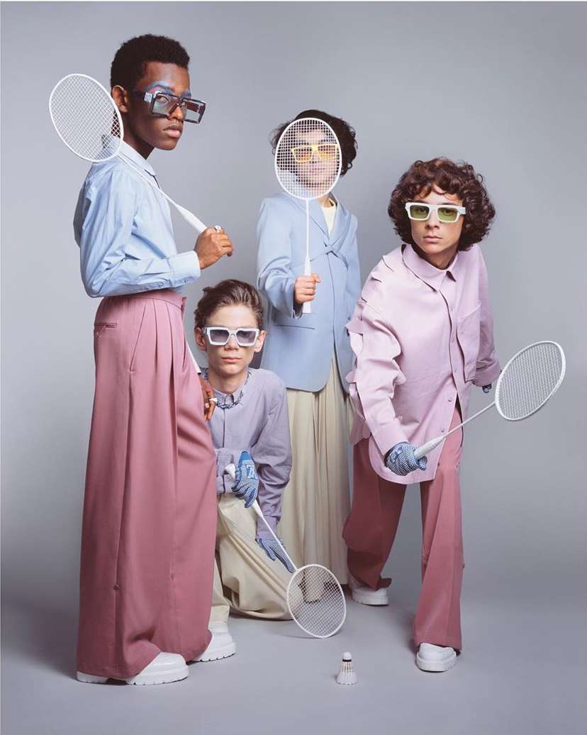 Four people in fashionable athletic attire pose with badminton racquets and a shuttlecock on a solid grey studio background.