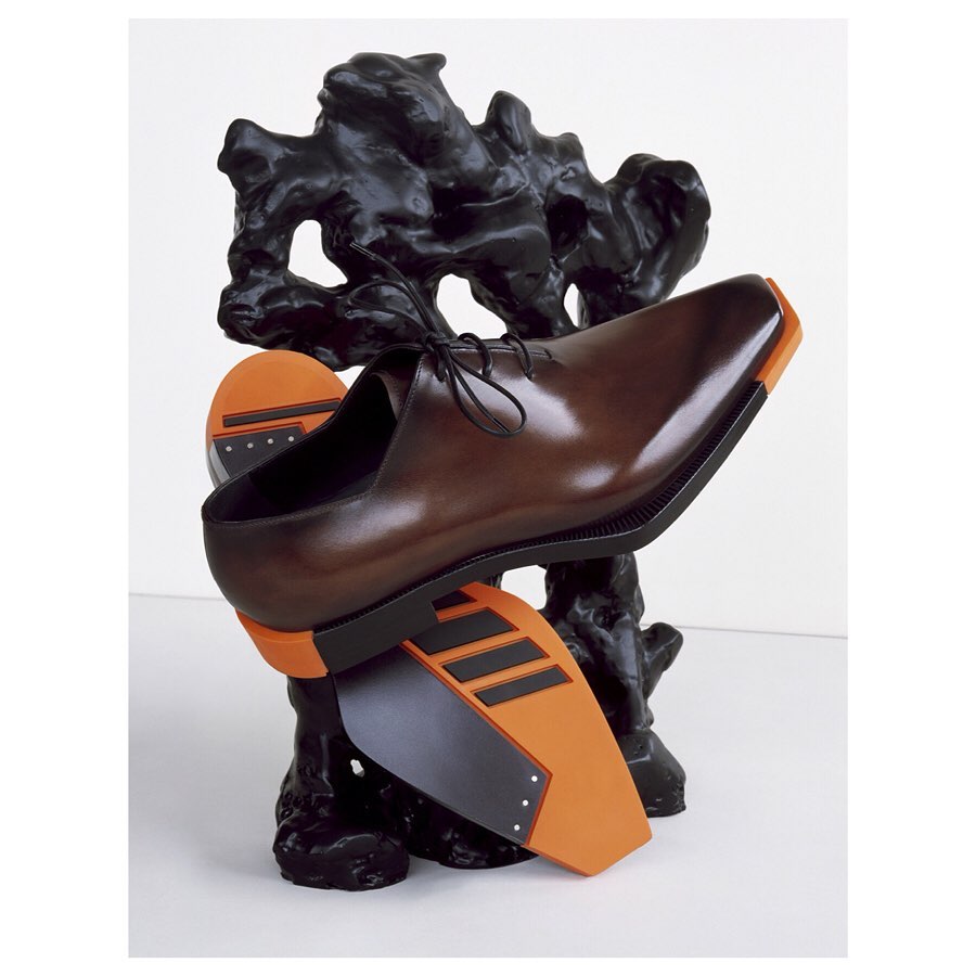 Studio shot of a brown leather dress shoe balanced atop an abstract black sculptural form with orange and black geometric platforms.