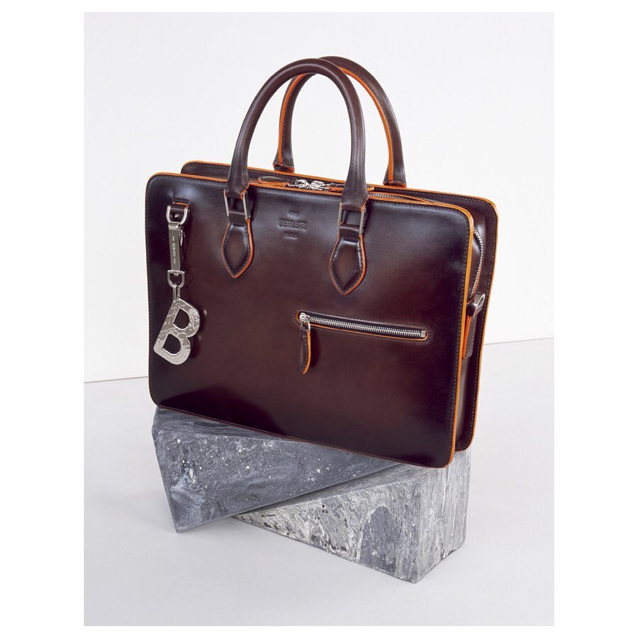Brown leather briefcase with silver hardware and 'B' charm, placed on grey marble blocks against a plain white background.