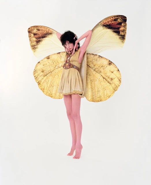 Young woman with dark hair poses in a tan dress with large butterfly wings on a white background, fashion forward, expressive.