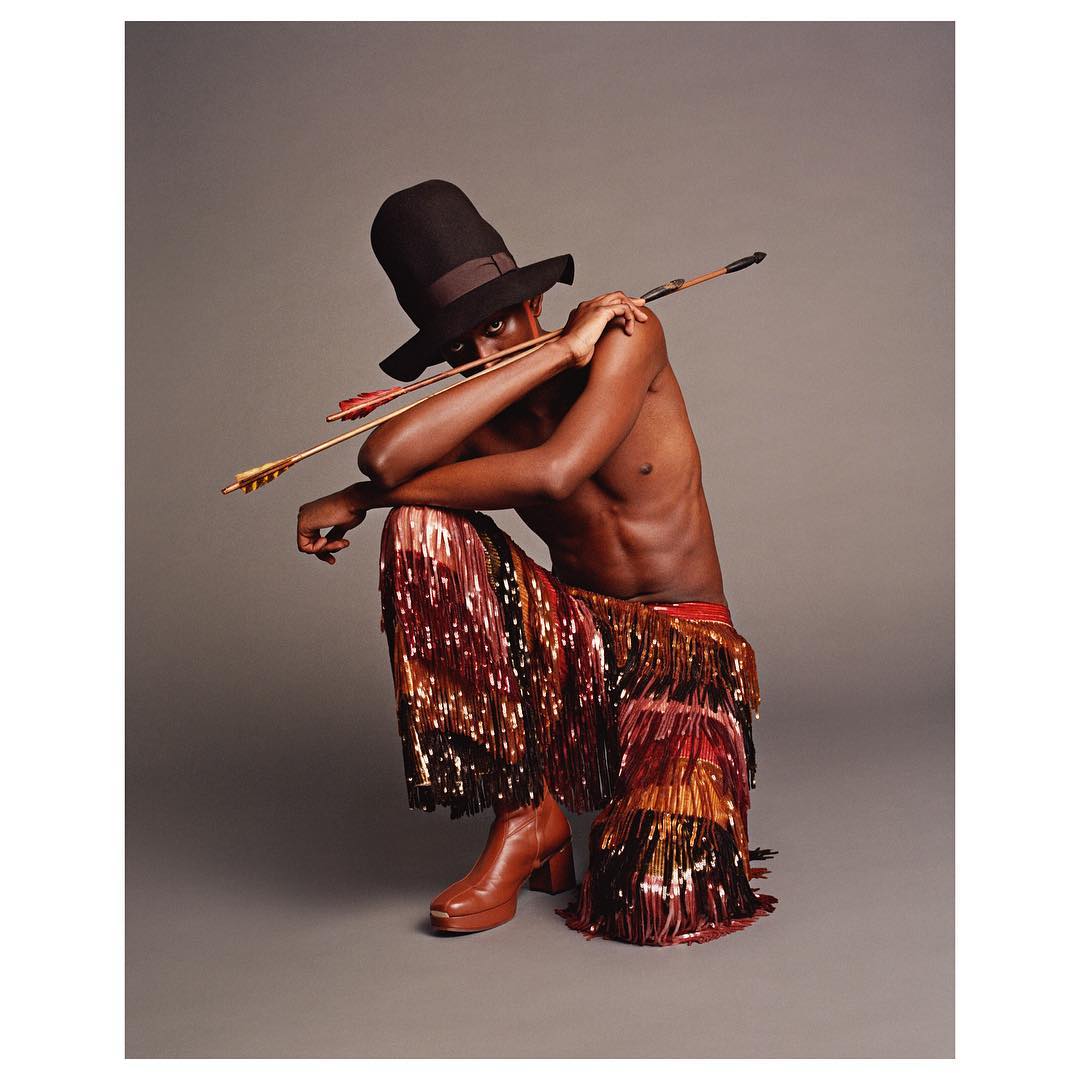 A slender Black man in an oversized black hat and fringed pants poses crouching with an arrow across his shoulders in a studio shot.