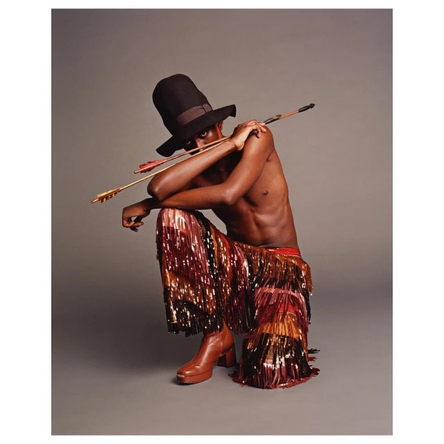 A slender Black man in an oversized black hat and fringed pants poses crouching with an arrow across his shoulders in a studio shot.
