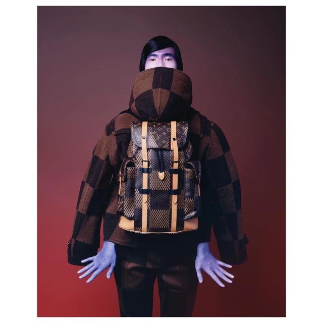 Fashion shot featuring an adult with painted blue skin wearing a brown checker pattern jacket and a Louis Vuitton backpack against a gradient background.