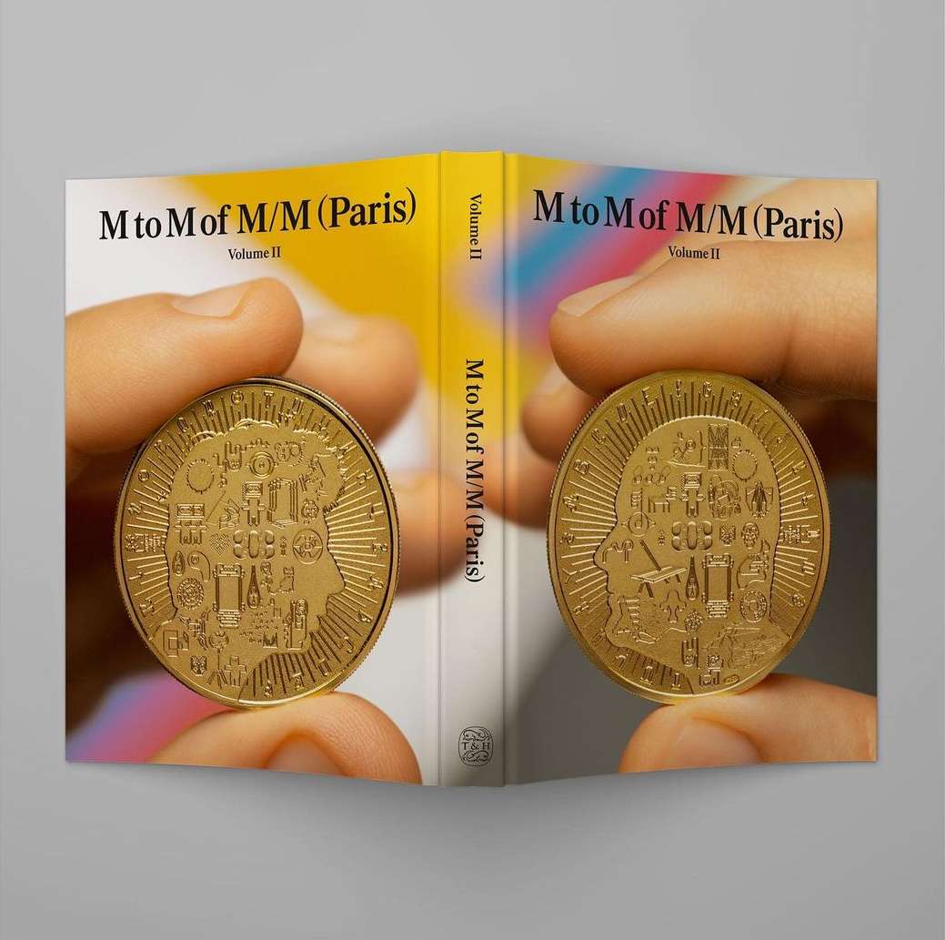 Book cover of M to M of M/M (Paris) Volume II held open, featuring two gold coins held in hands against the cover's yellow and white design.