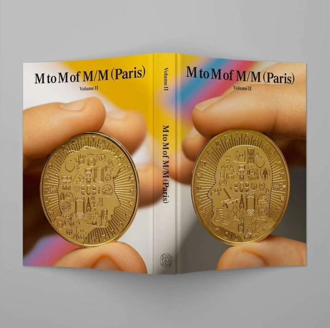 Book cover of M to M of M/M (Paris) Volume II held open, featuring two gold coins held in hands against the cover's yellow and white design.