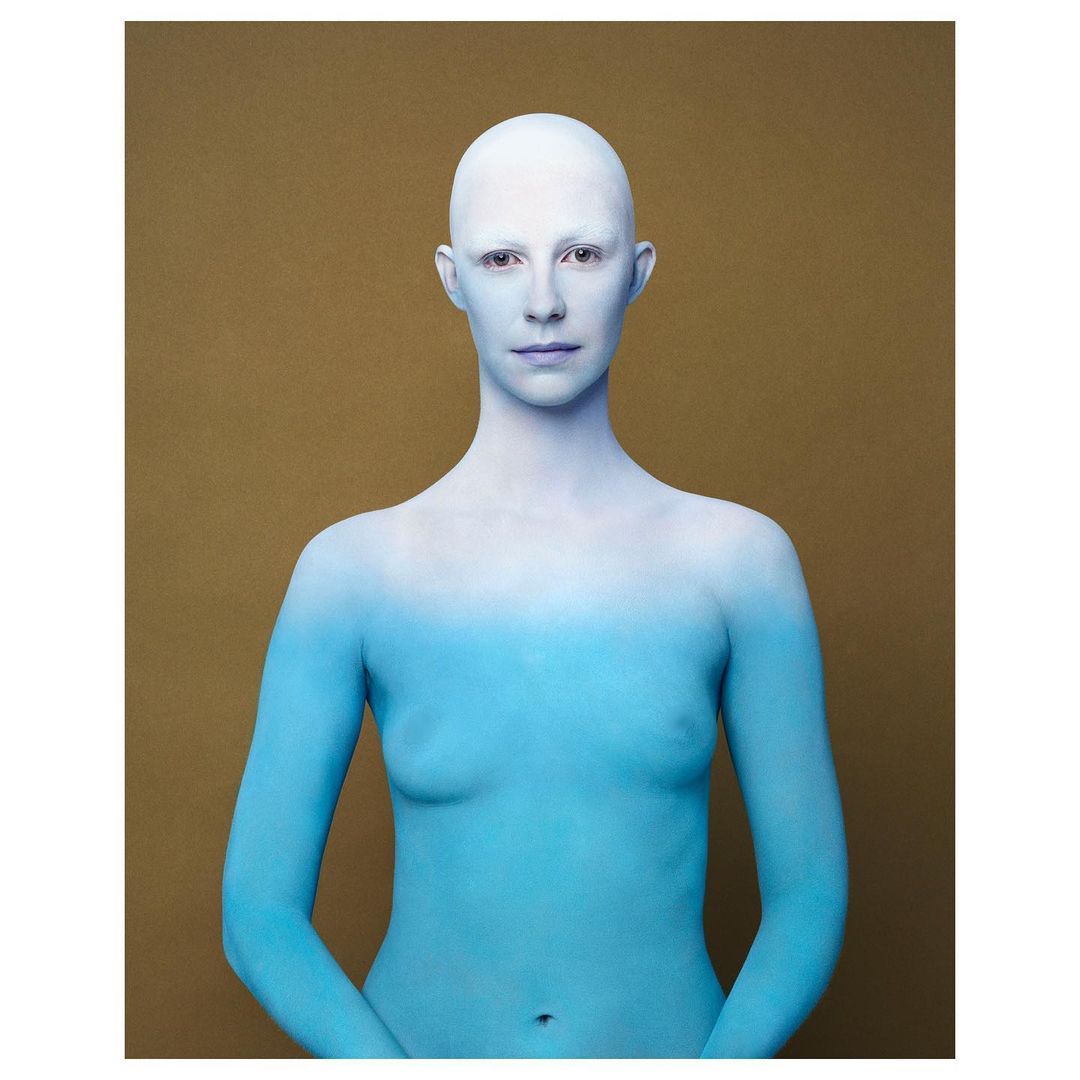 Close-up portrait of an adult woman with bald head and body paint in light blue and white against a brown background in a studio setting.