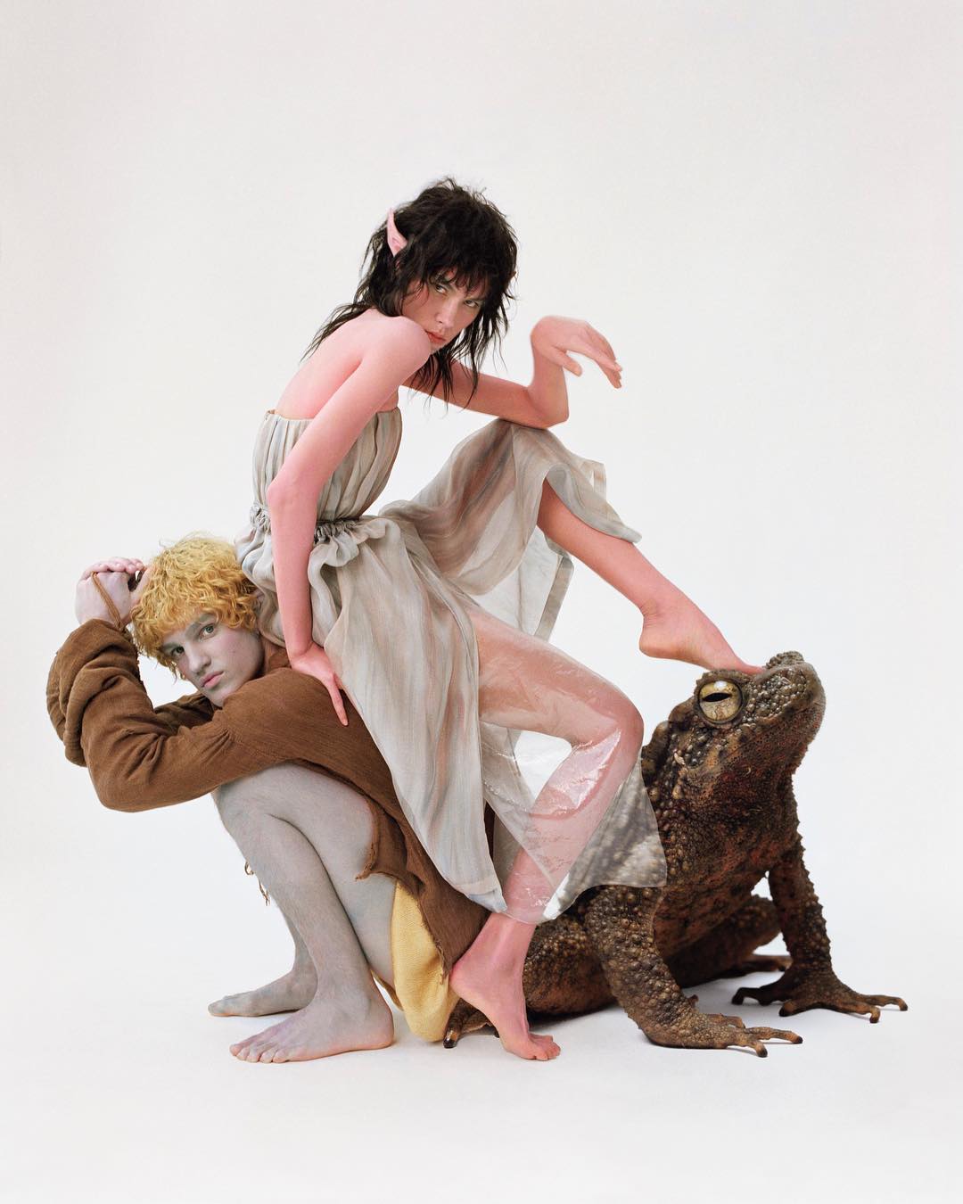 Two figures in fantasy-inspired fashion with elf ears, one kneeling and supporting the other, plus a large toad on a white backdrop.