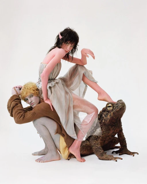 Two figures in fantasy-inspired fashion with elf ears, one kneeling and supporting the other, plus a large toad on a white backdrop.