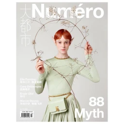 Model with red hair and prosthetic elf ears holding a twig circle wearing a pale green long-sleeve top against a grey background.