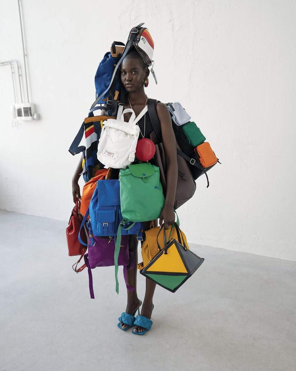 Fashionable woman poses indoors surrounded by colorful backpacks, wearing strappy high heels in a brightly lit studio setting.
