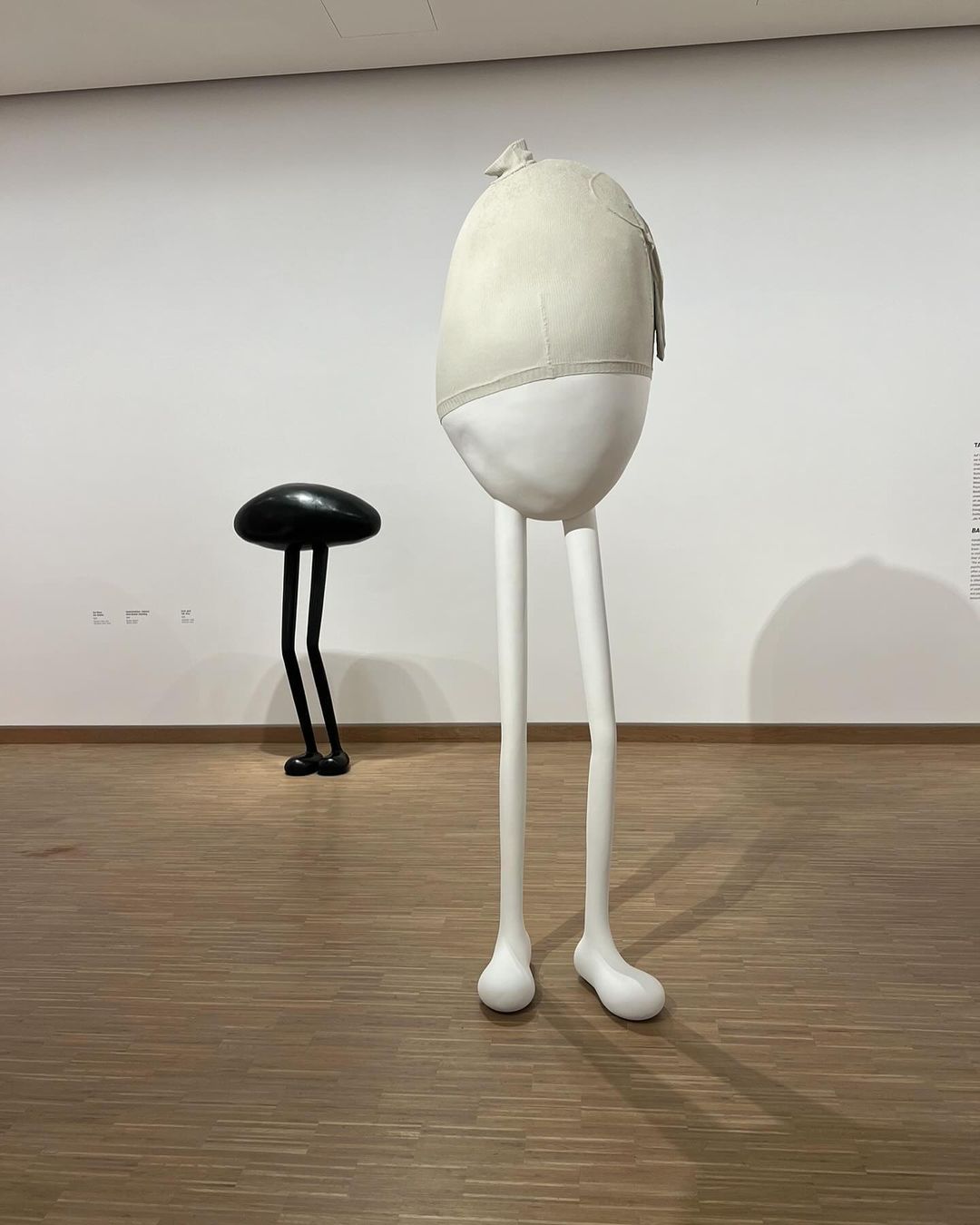 Art installation featuring a large, egg-shaped sculpture with stick-like legs and a smaller, dark, mushroom-shaped sculpture on a wood floor.