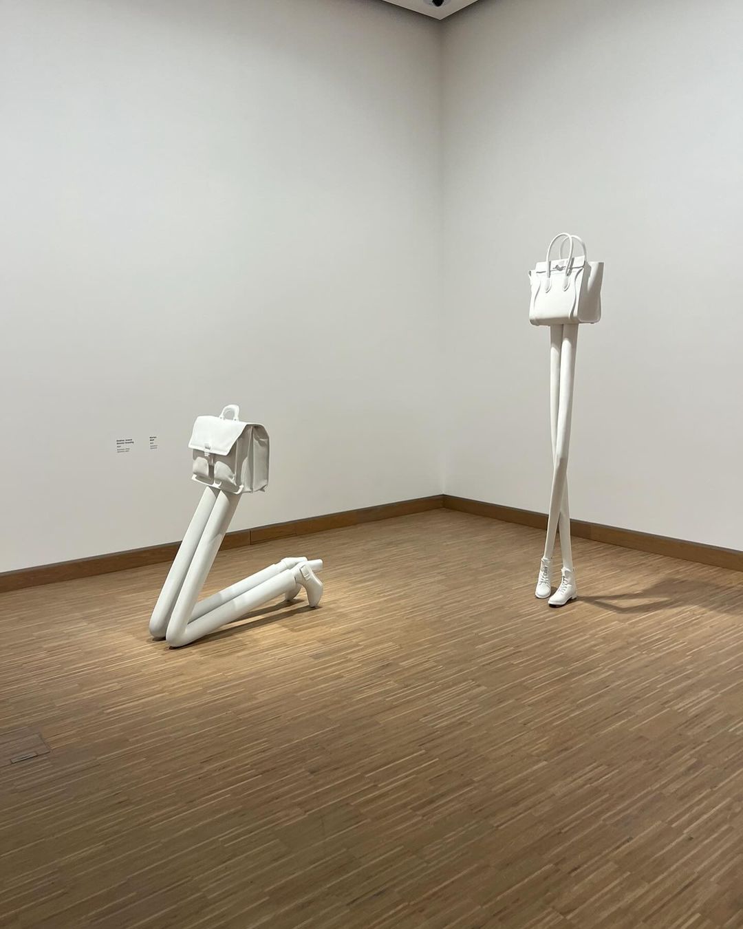 Contemporary art exhibit: two white handbag sculptures with leg-like bases stand in an art gallery with light wood floors and white walls.