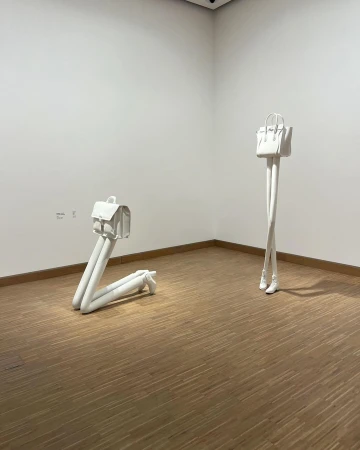 Contemporary art exhibit: two white handbag sculptures with leg-like bases stand in an art gallery with light wood floors and white walls.