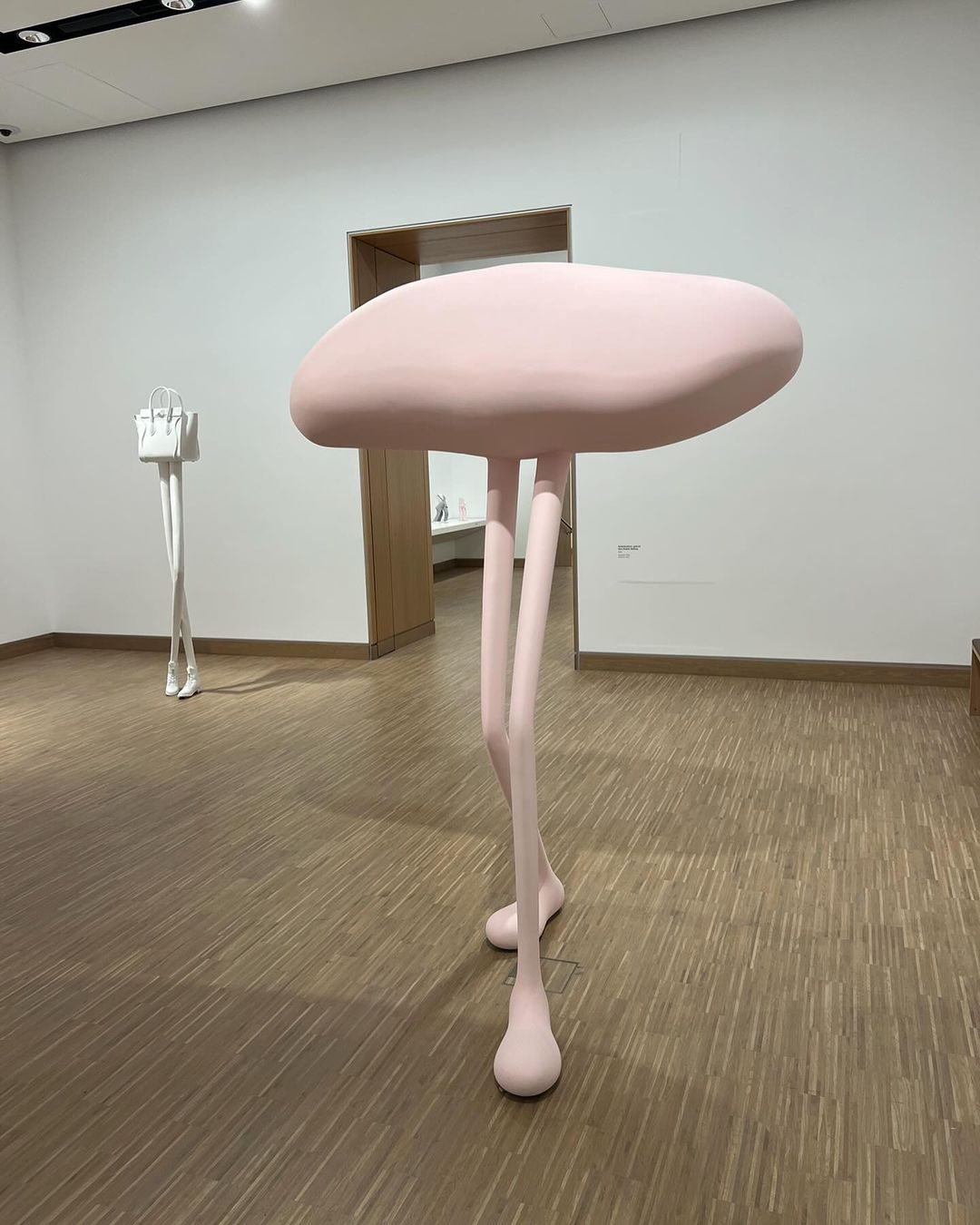 Sculpture of an abstract figure with bag in an art gallery with white walls and wooden floor. The figure is pink with thin legs.